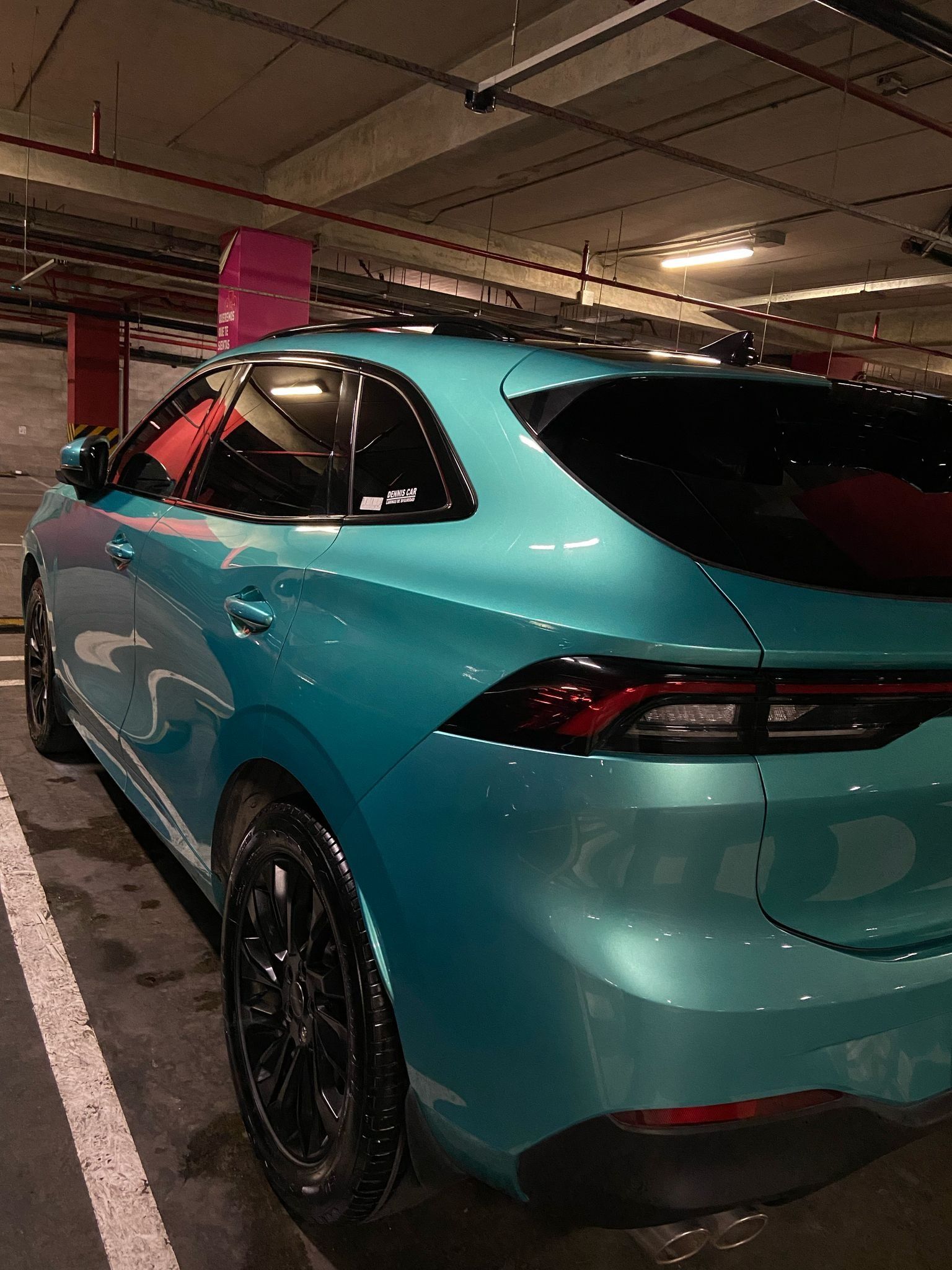 Un coche verde azulado aparcado en un aparcamiento oscuro. Las llantas negras contrastan con la pintura metalizada, junto a una línea blanca de aparcamiento.