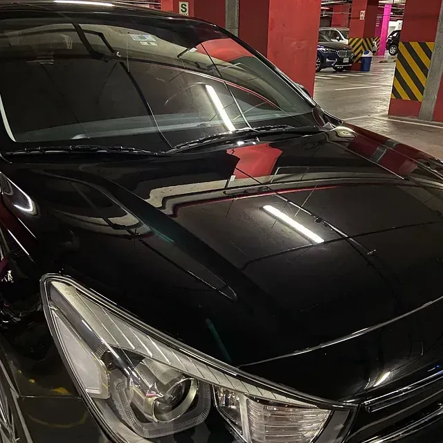 Coche negro aparcado en un aparcamiento con luces de techo reflejándose en el capó.