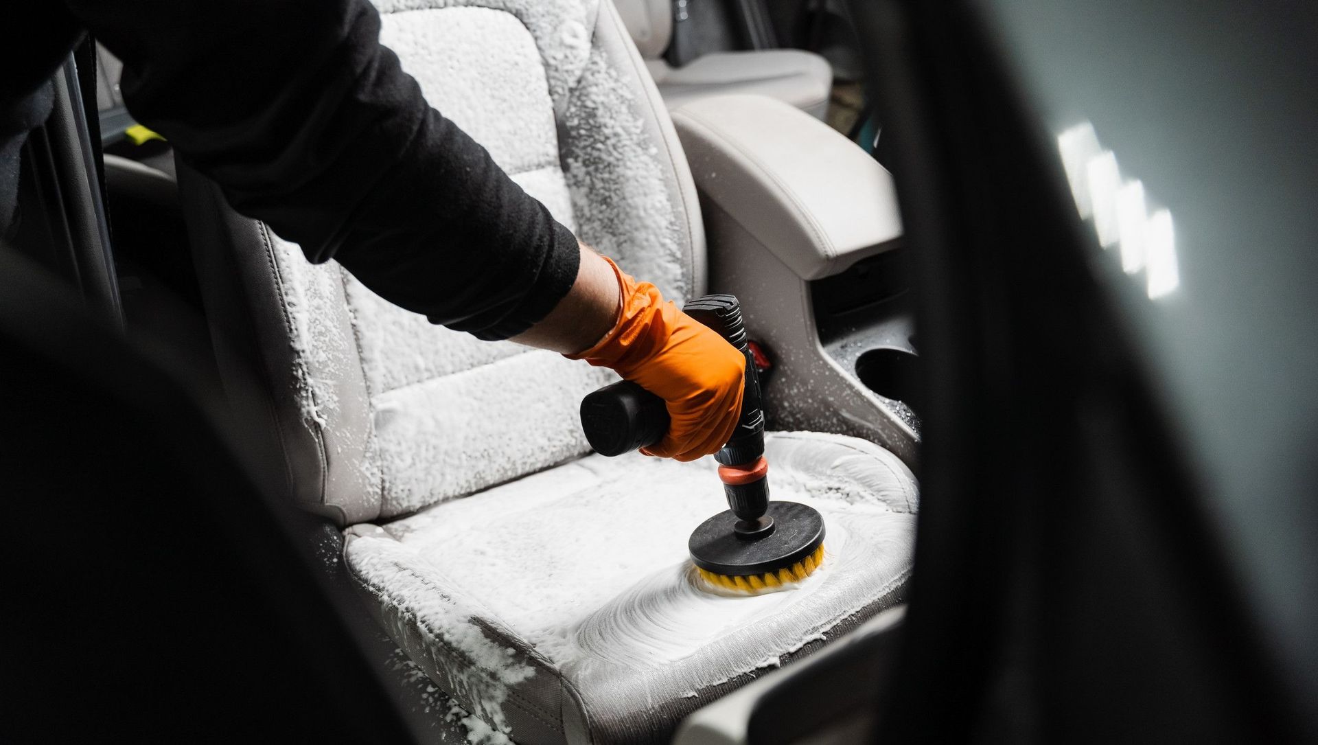 Persona con guantes naranjas usando un taladro eléctrico con cepillo para fregar el asiento de un coche. Dentro de un coche.