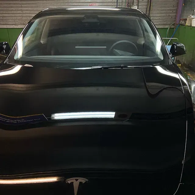Coche Tesla de color negro, vista frontal, acabado brillante, estacionado en un espacio interior, con luces superiores brillantes reflejándose en el capó.