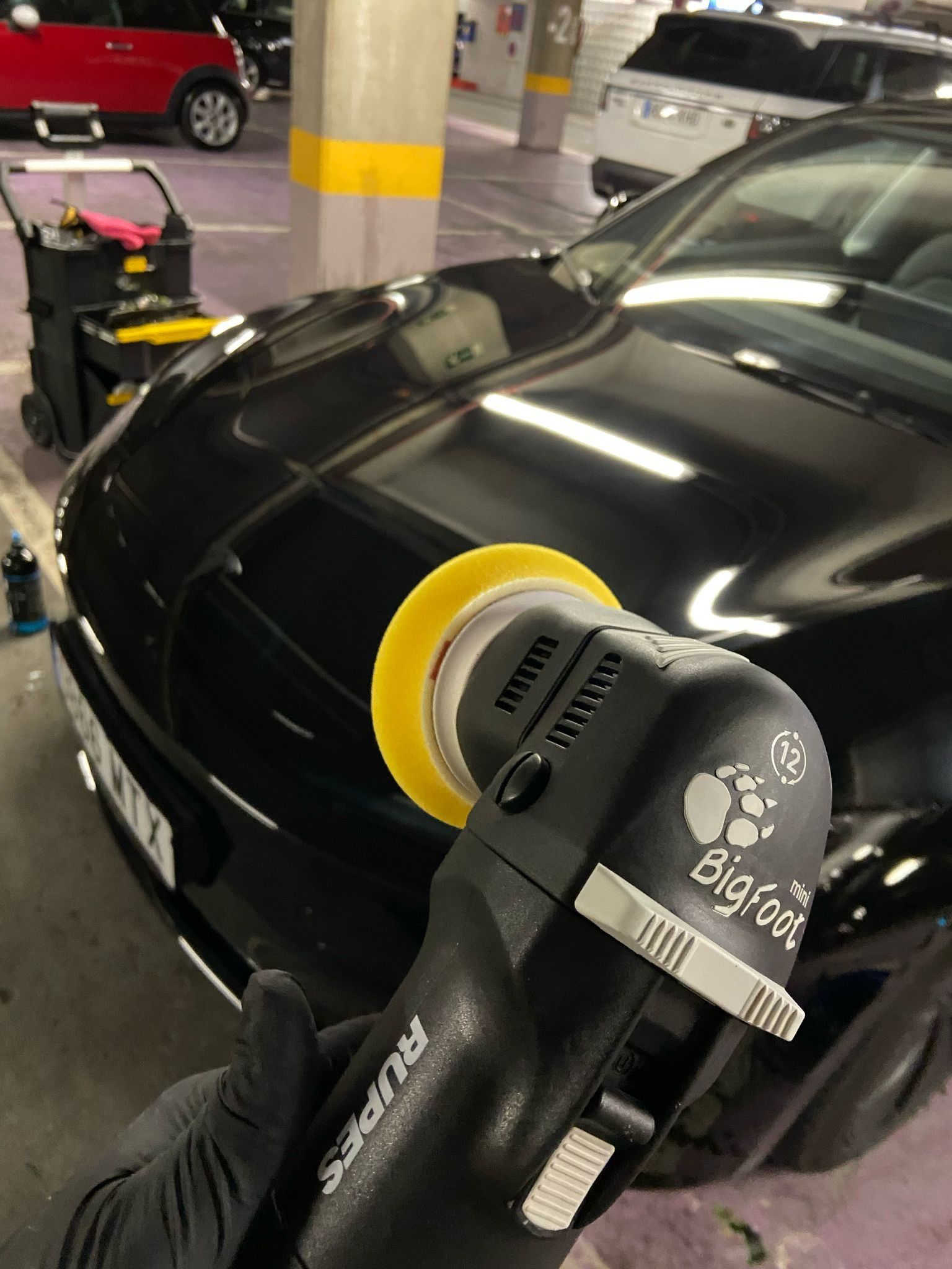 Una persona con guantes negros utiliza un pulidor negro y amarillo sobre un automóvil negro brillante en un estacionamiento.