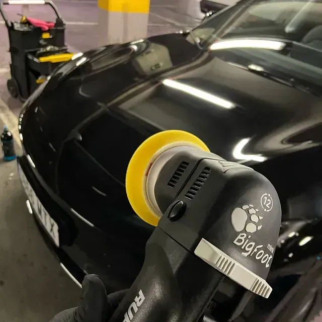Una persona puliendo un coche negro con una pulidora orbital con almohadillas amarillas en un garaje muy iluminado.