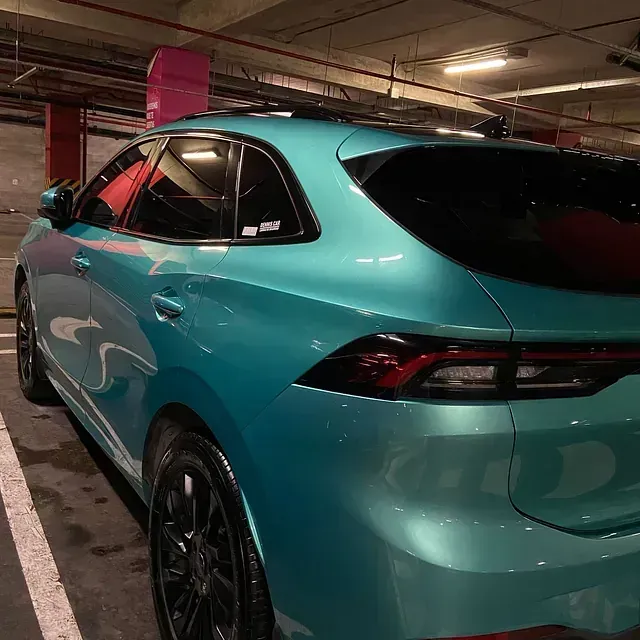 Una camioneta verde azulado estacionada en un estacionamiento con poca luz. El auto tiene llantas negras y ventanas tintadas.