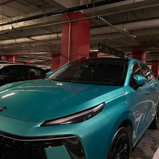 Un coche verde azulado aparcado en un aparcamiento con columnas rojas. El coche está en ángulo hacia la cámara.