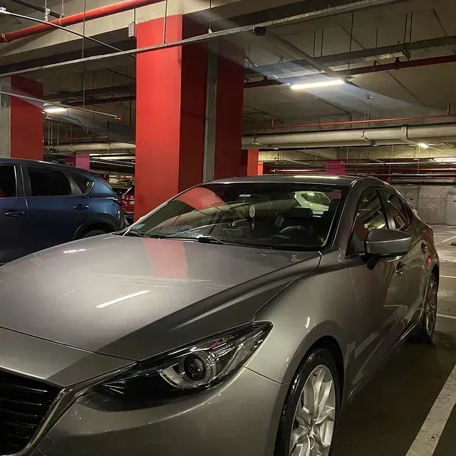 Un sedán Mazda plateado estacionado en un estacionamiento de varios niveles con columnas de soporte rojas y tubos elevados.