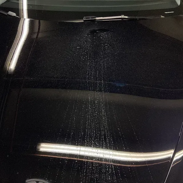 Capó de coche negro con agua saliendo a chorro hacia arriba, reflejando la luz.