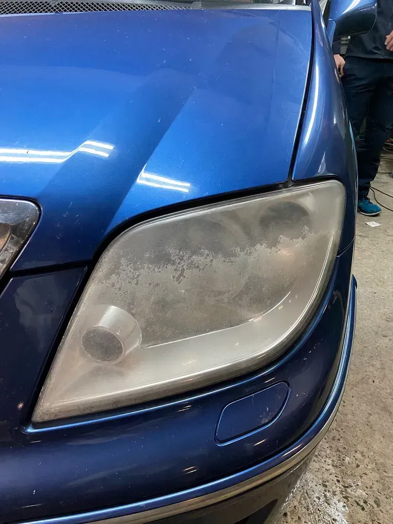 Coche azul con un faro nublado, al fondo se ven las piernas de una persona.