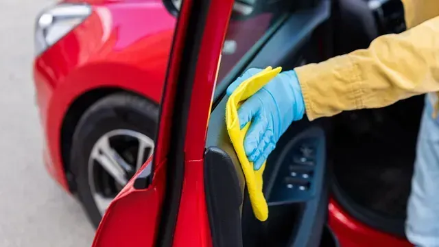 Una persona que lleva guantes azules y una camisa amarilla limpia la puerta de un coche rojo con un paño amarillo.