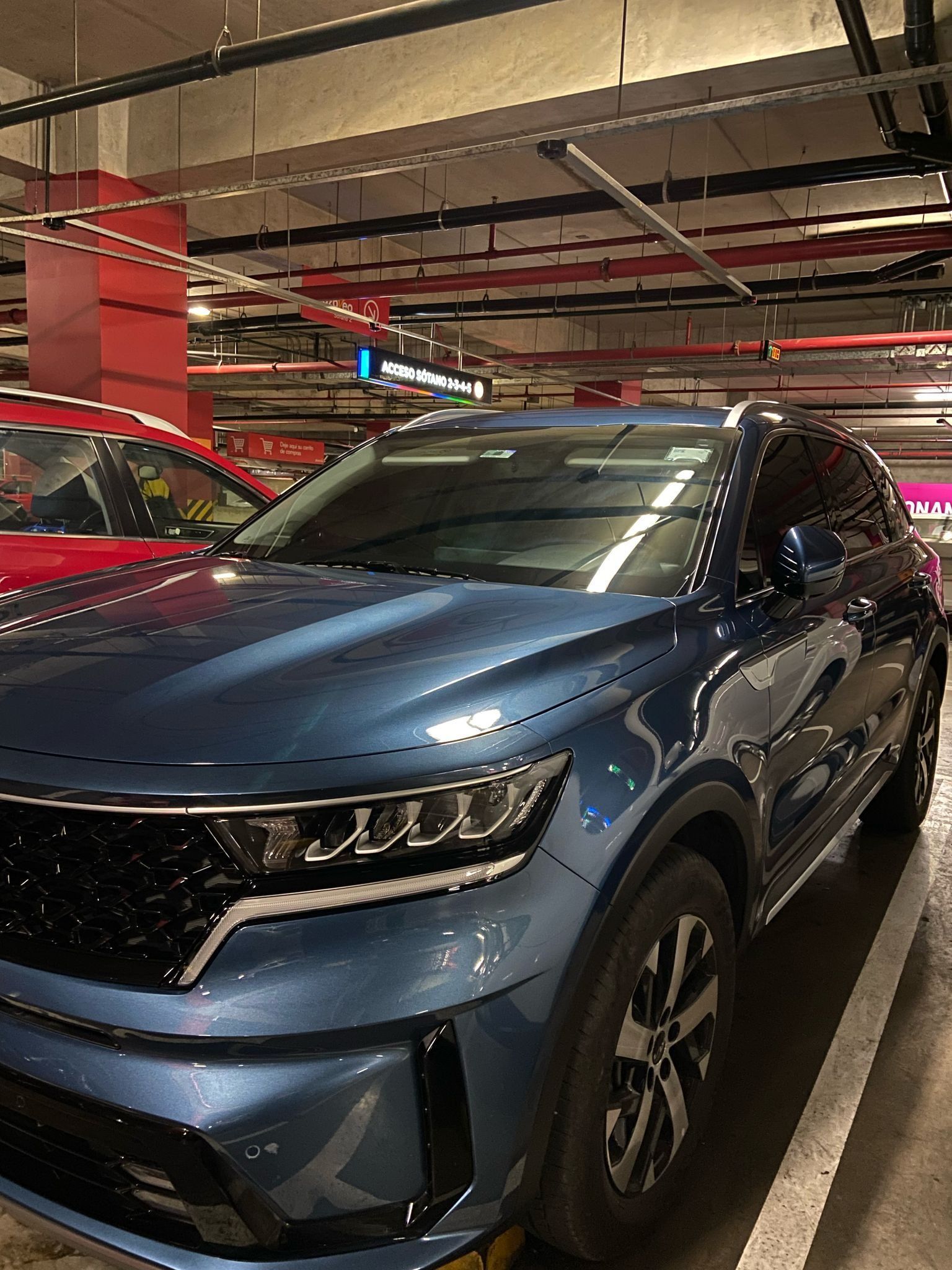 SUV Kia Sorento azul estacionada en un estacionamiento interior. Primer plano del frontal. Columnas de soporte rojas y tubos superiores visibles.