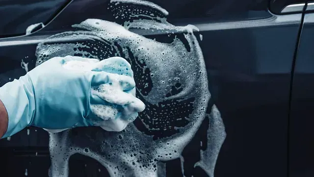 Mano enguantada lavando un coche oscuro con una esponja jabonosa; toma de primer plano.