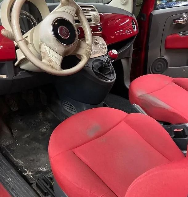 Interior de un Fiat 500 rojo, con asientos rojos desgastados, volante color blanquecino y consola negra.