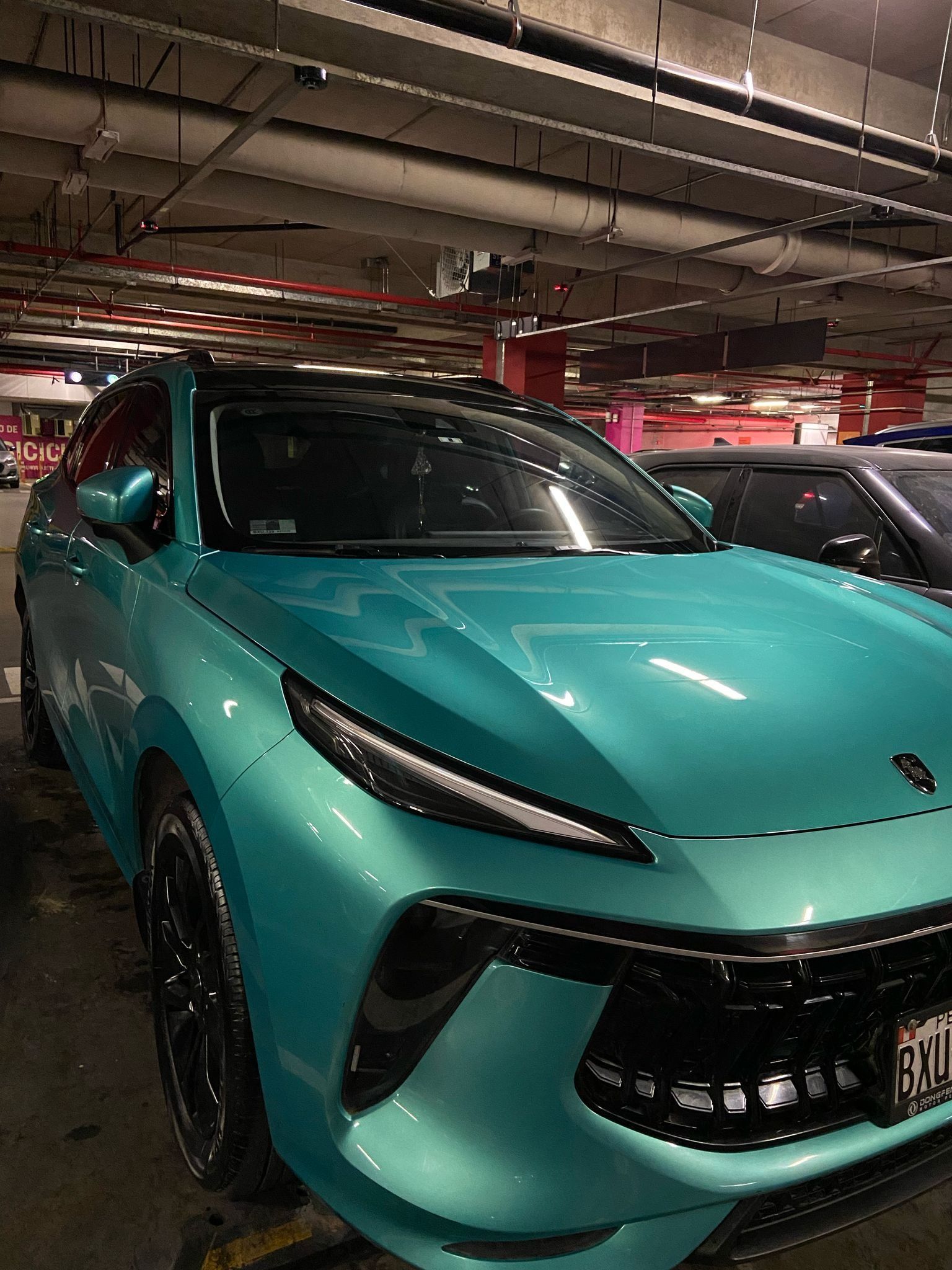 Una camioneta verde azulado estacionada en un estacionamiento, con llantas y techo negros. El ambiente es interior, con luces fluorescentes en el techo.