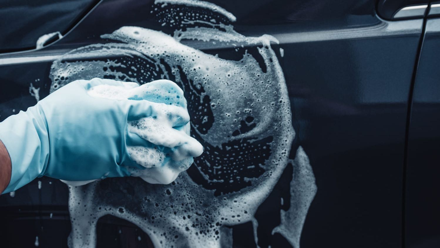 Una persona que lleva un guante azul lava un coche de color oscuro con una esponja jabonosa.