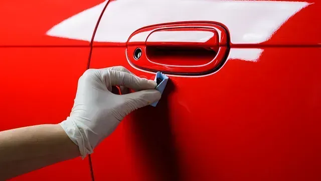 Mano enguantada que utiliza una barra de arcilla azul para limpiar la puerta roja de un automóvil cerca de la manija.