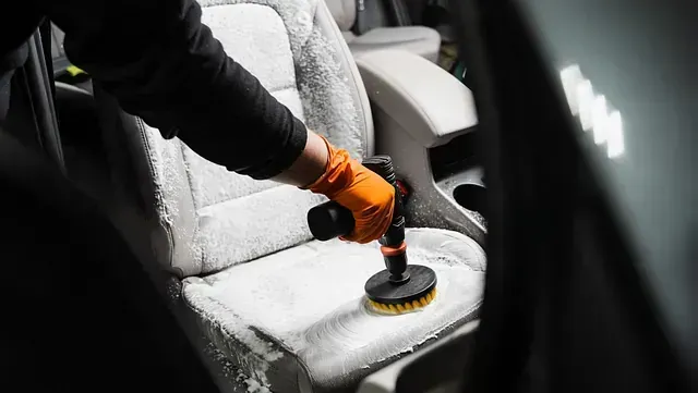 Una persona que lleva un guante naranja utiliza un accesorio de cepillo en una herramienta eléctrica para limpiar el asiento de un automóvil.