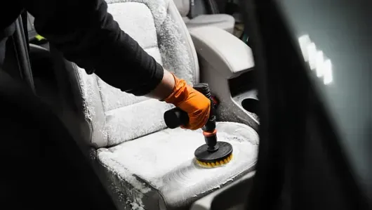 Una persona que lleva un guante naranja utiliza un accesorio de cepillo en una herramienta eléctrica para limpiar el asiento de un automóvil.