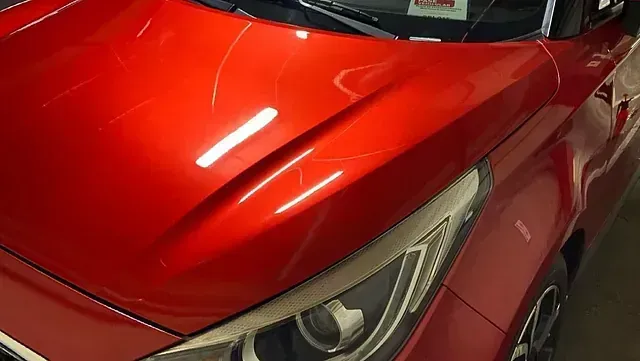 Capó de coche rojo con arañazos que reflejan las luces del techo.