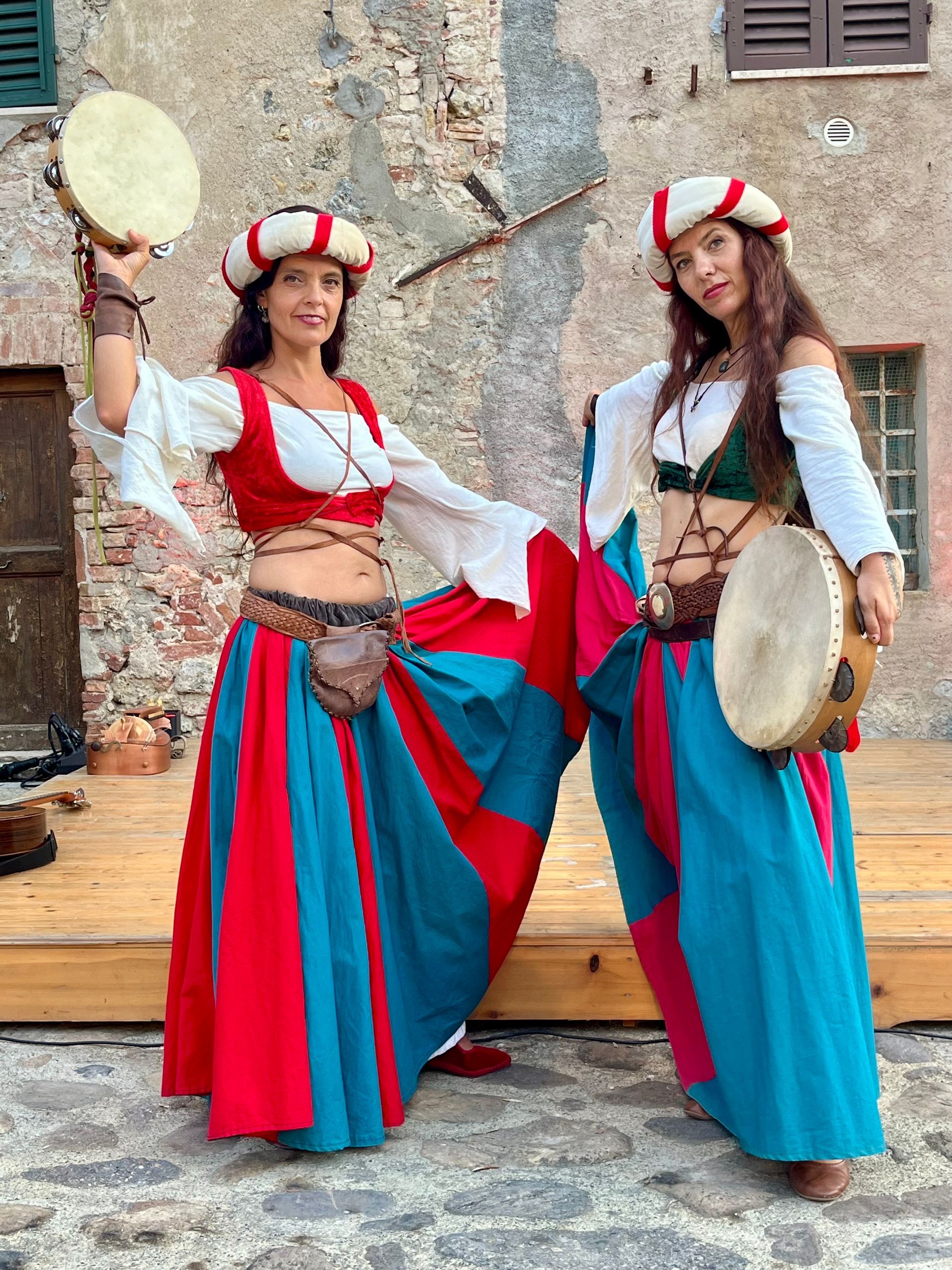 Dos mujeres con trajes renacentistas con panderetas. Al aire libre, con un edificio como telón de fondo, faldas coloridas.