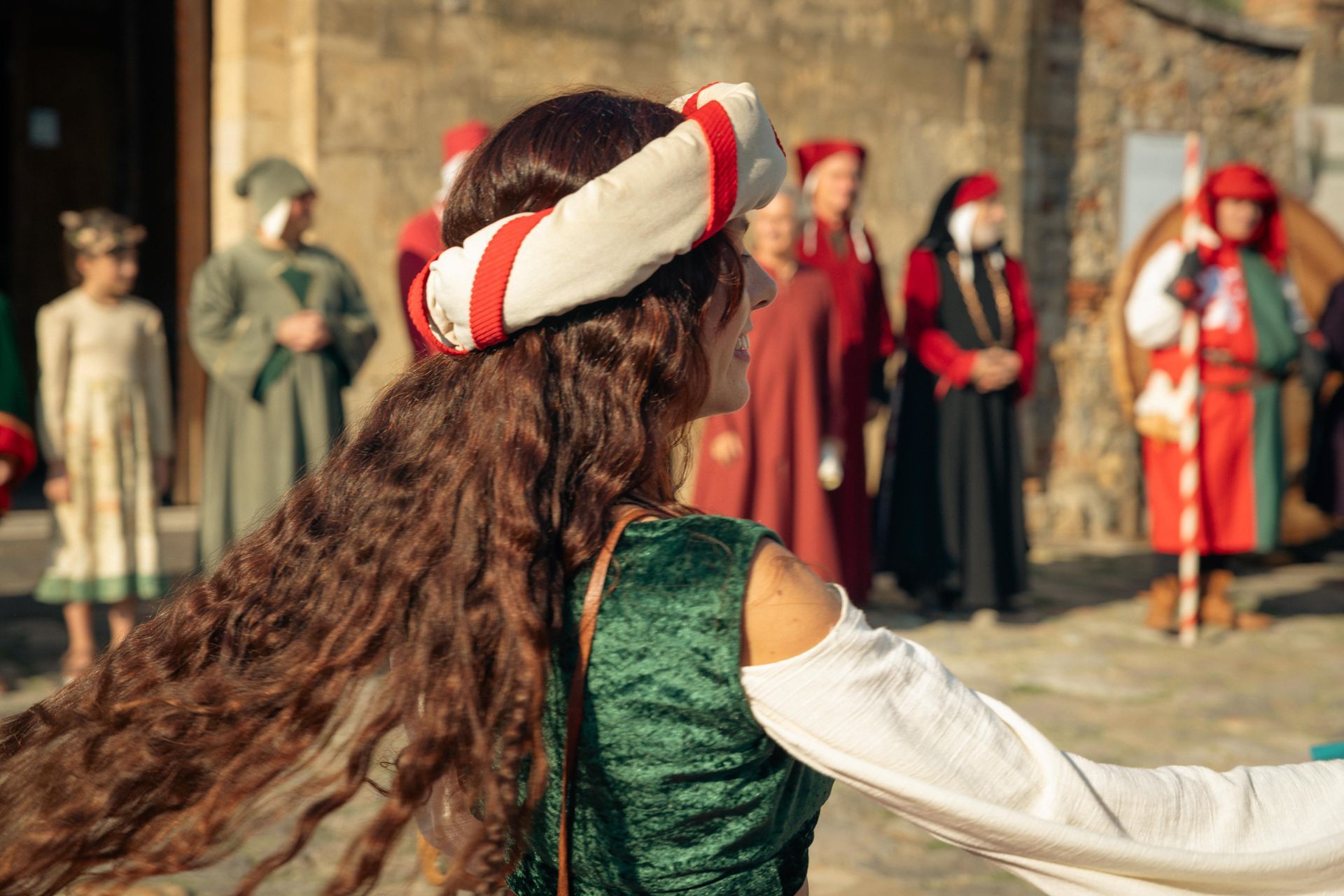 Mujer con traje medieval, pasando por un desfile, cabello largo castaño, tocado blanco y rojo, otras personas disfrazadas.