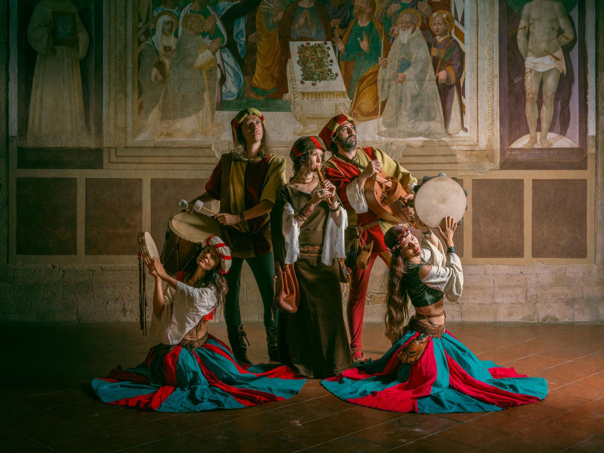Músicos y bailarines medievales con trajes coloridos tocan instrumentos frente a un fresco.