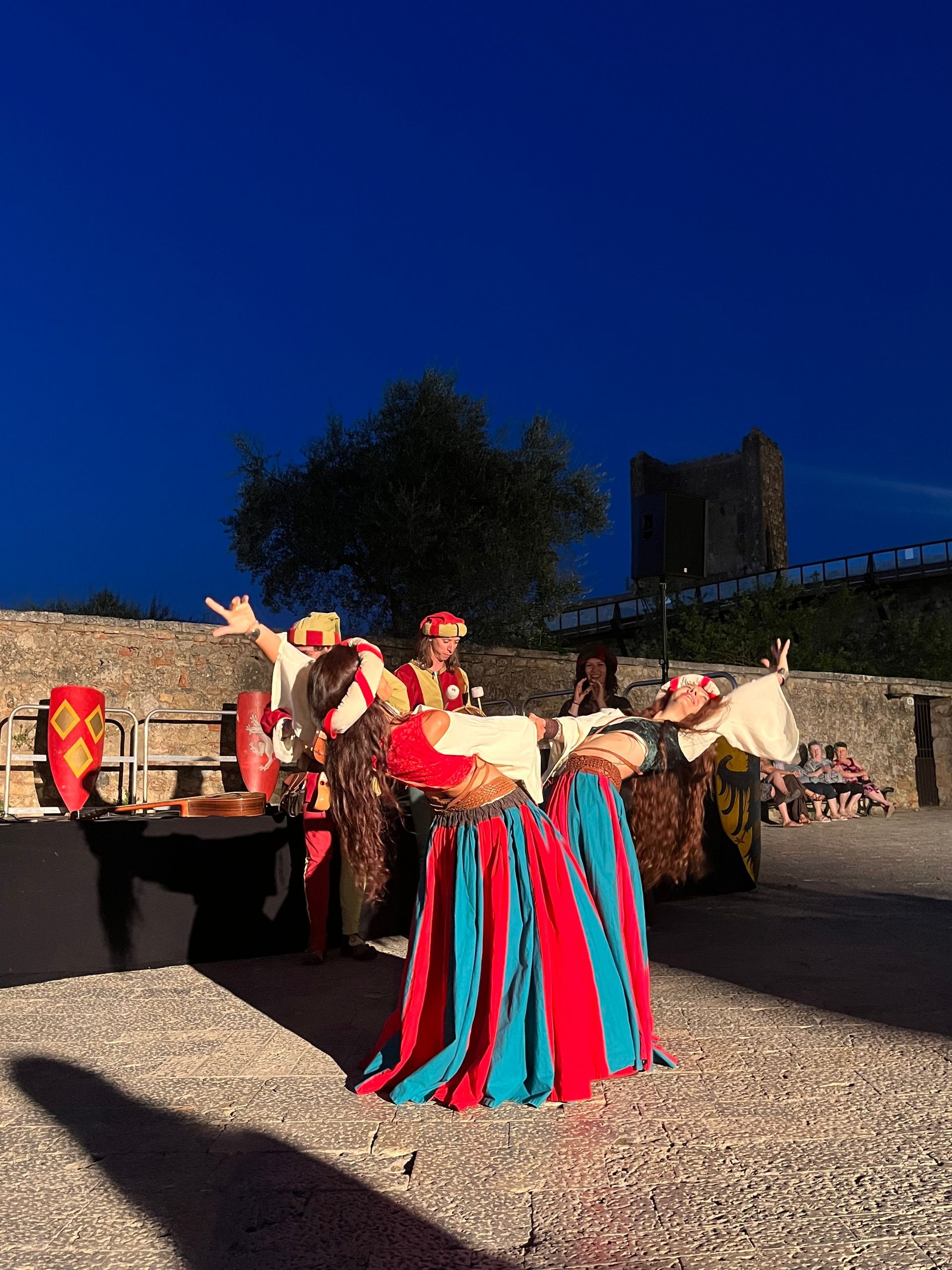 Artistas con trajes medievales en un evento al aire libre; una mujer baila, otros observan; fondo de piedra y cielo oscuro.