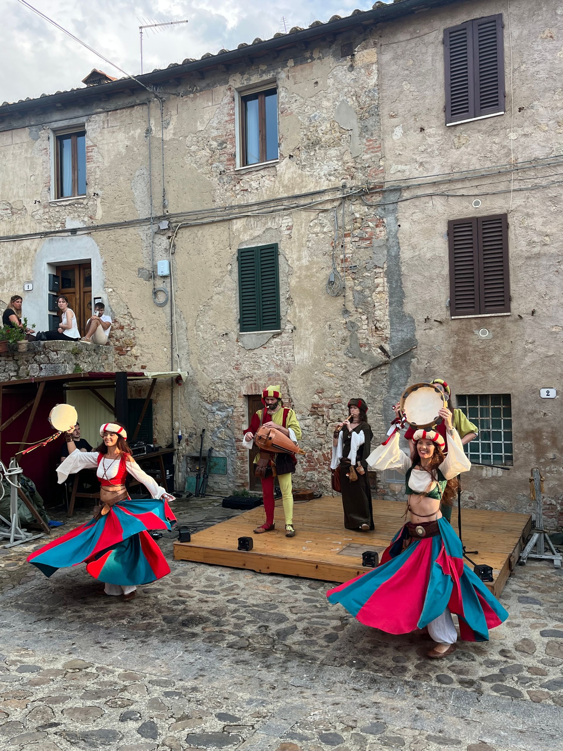 Artistas medievales en el escenario frente a un edificio de piedra; bailarines con faldas rojas y azules giran.