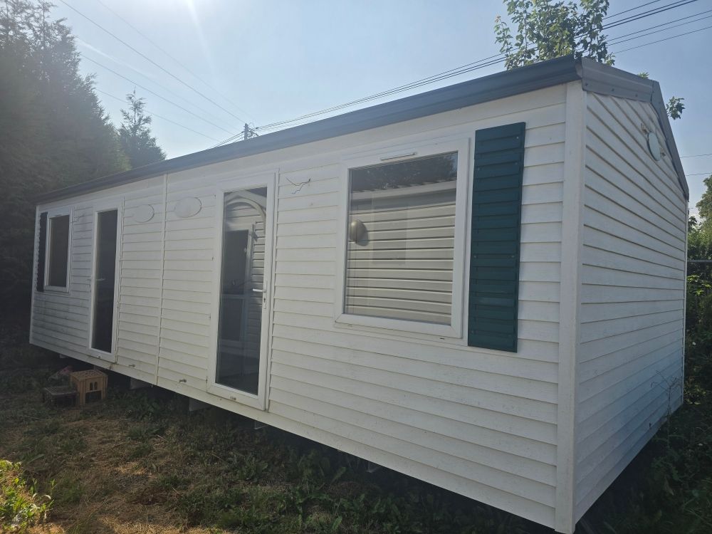 Petite caravane blanche aux volets sombres et à la porte ouverte, installée sur un terrain herbeux sous un soleil éclatant.