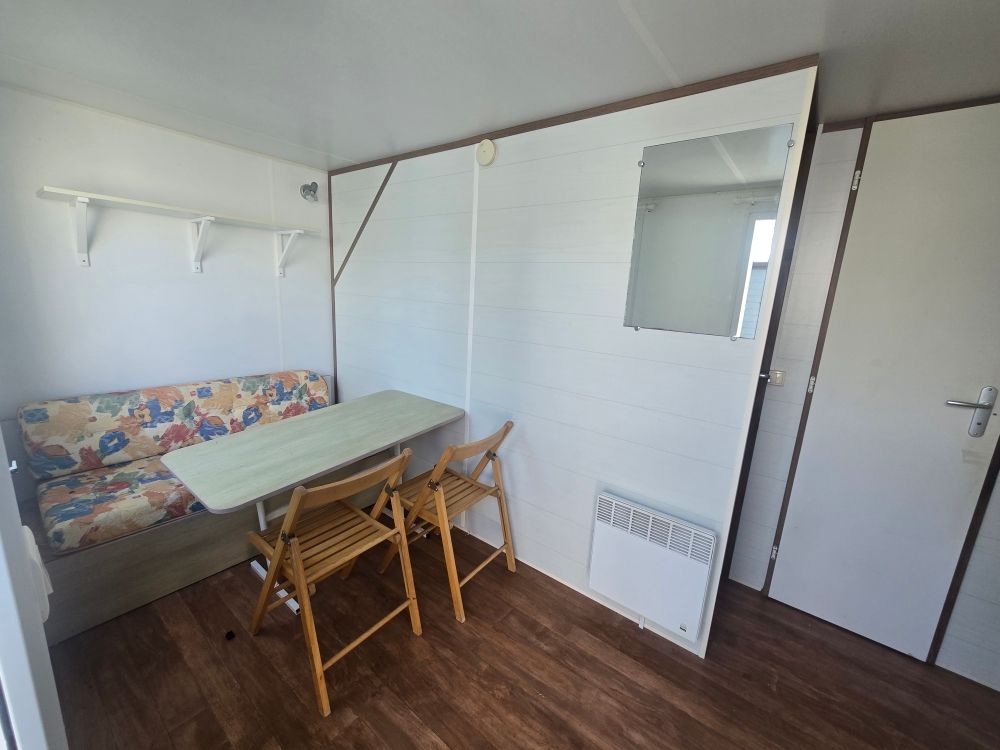 Petite pièce avec une table, des chaises pliantes, un banc, des étagères murales, un miroir et une porte blanche sur un sol en bois.