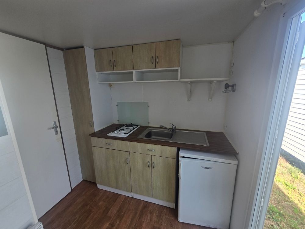 Petite kitchenette avec placards en bois, évier, plaque de cuisson et mini-frigo à côté d'une fenêtre.