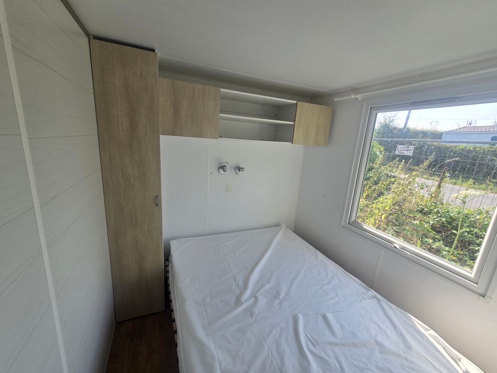 Petite chambre avec un lit, des armoires en bois et une grande fenêtre donnant sur la verdure.