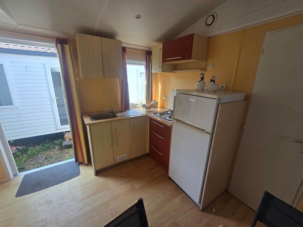 Petite cuisine avec réfrigérateur blanc, placards en bois, évier et porte ouverte sur l'extérieur.