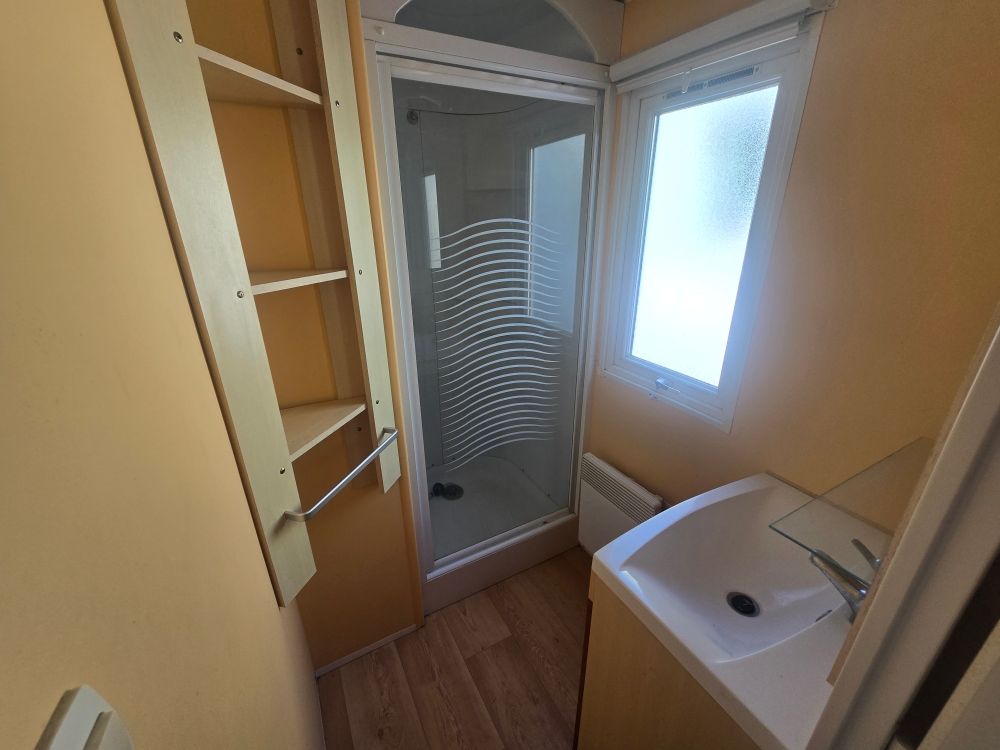 Petite salle de bains avec cabine de douche, lavabo et fenêtre, murs beiges et sol imitation bois.