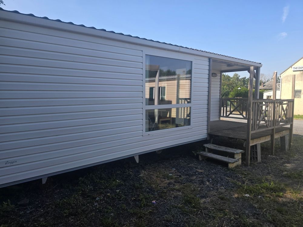Mobil-home blanc avec une petite véranda en bois et quelques marches, sur un terrain gravillonné sous un ciel bleu.