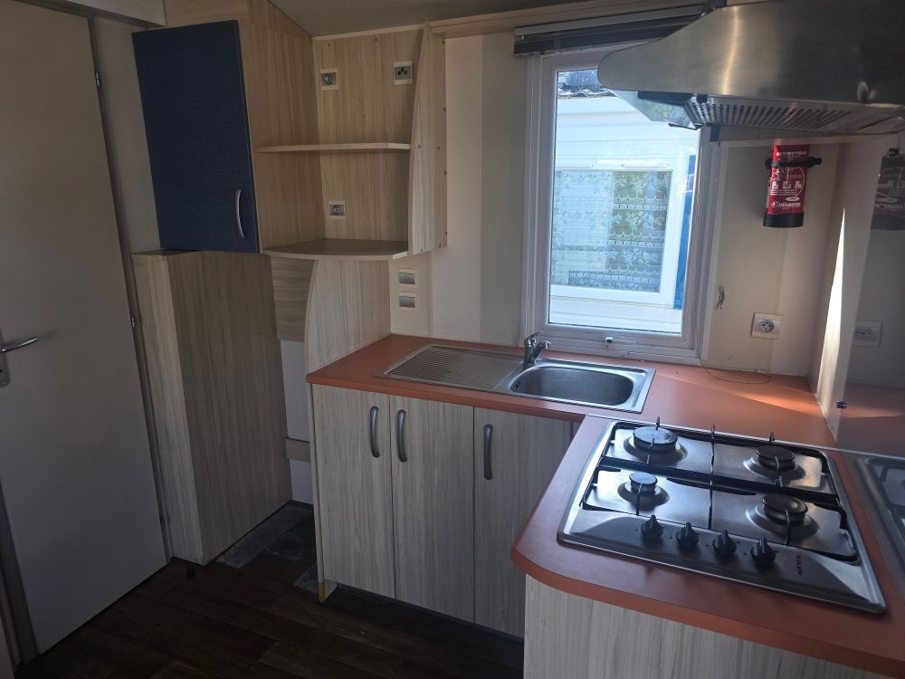 Cuisine compacte avec placards en bois, évier sous la fenêtre et plaque de cuisson à gaz sur un plan de travail rouge.