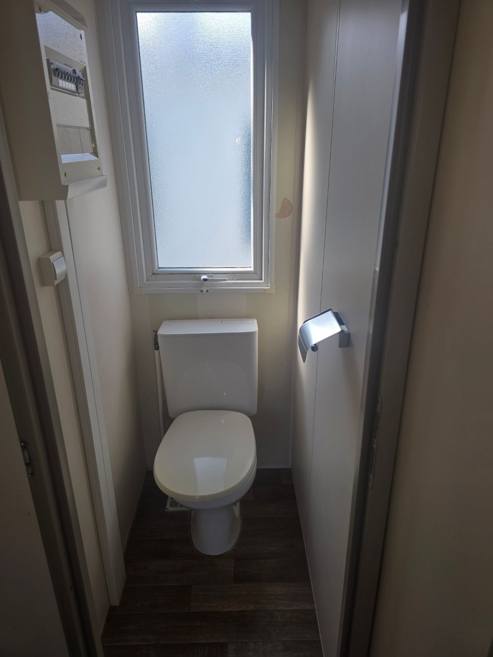 Salle de bains étroite avec toilettes blanches, fenêtre dépolie et porte-papier toilette mural.
