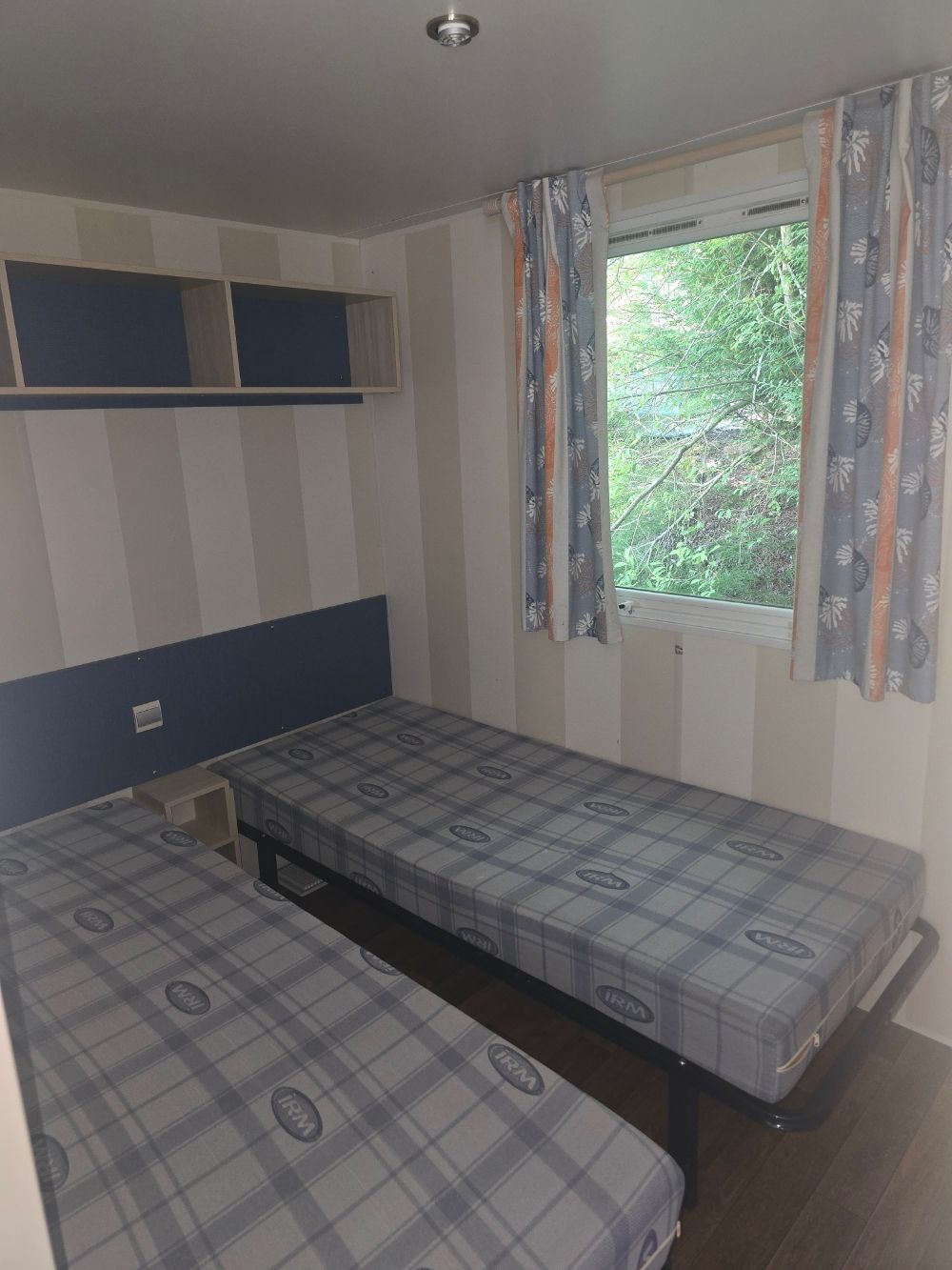 Petite chambre avec deux lits simples, des touches de bleu et une fenêtre avec des rideaux à motifs.