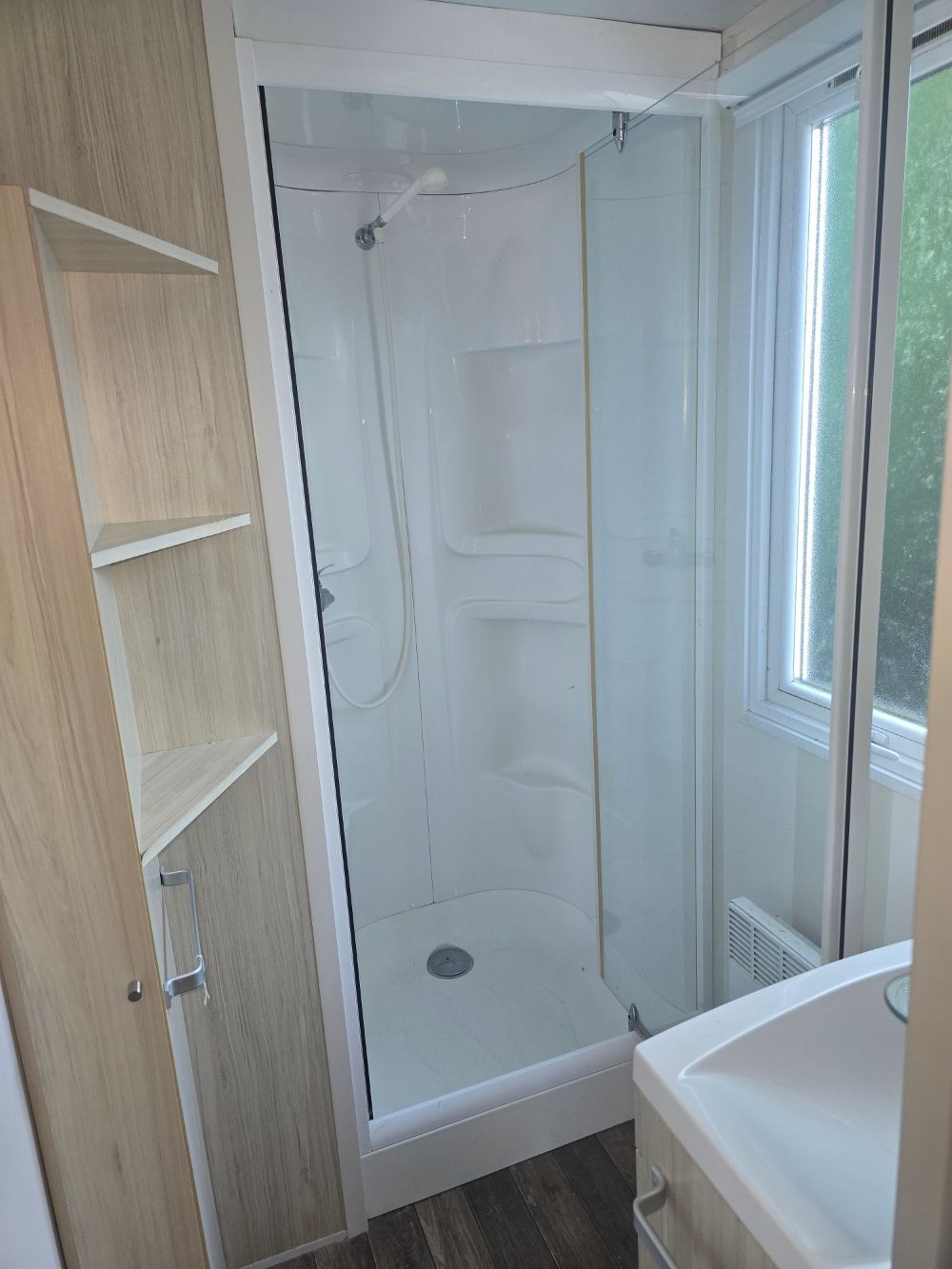 Cabine de douche étroite avec murs blancs et porte vitrée à côté d'une fenêtre et étagères en bois intégrées.