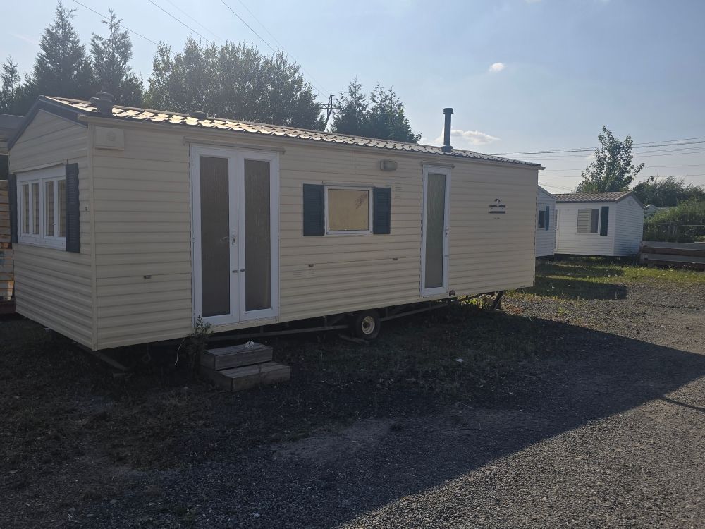 Petite maison mobile beige aux finitions blanches, sur un terrain de gravier sous un ciel radieux.