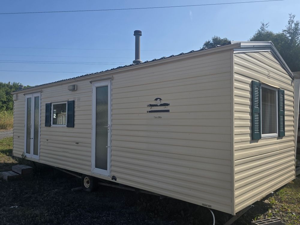 Petite maison préfabriquée beige sur roues, avec des volets verts et une porte blanche, sous un ciel bleu.