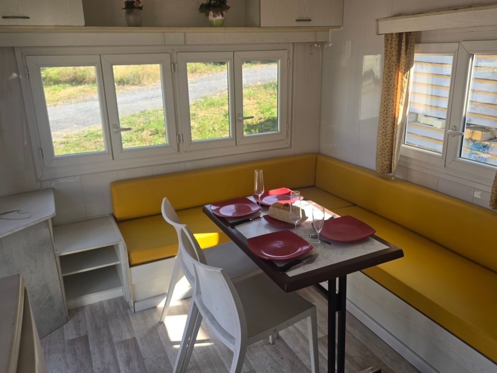 Coin repas lumineux avec banquettes blanches et jaunes, petite table dressée avec des assiettes rouges et grandes fenêtres.