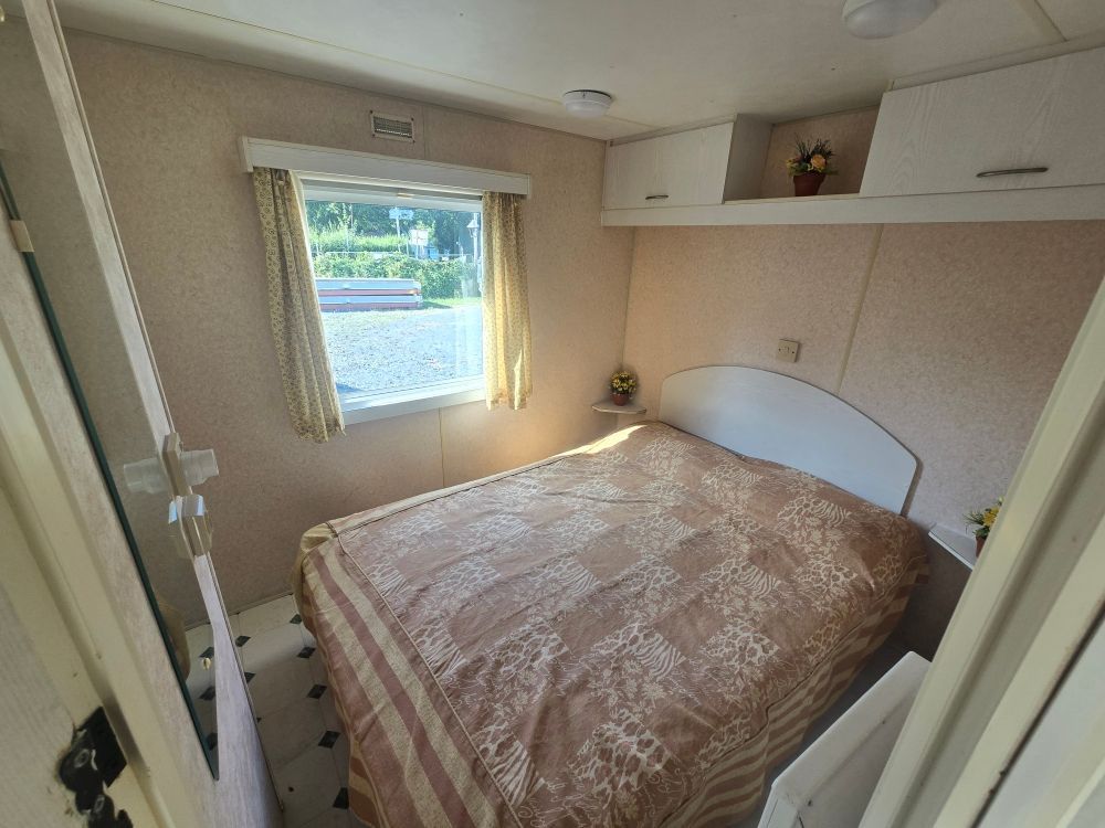 Petite chambre avec un lit, une fenêtre, des murs beiges et des placards intégrés.
