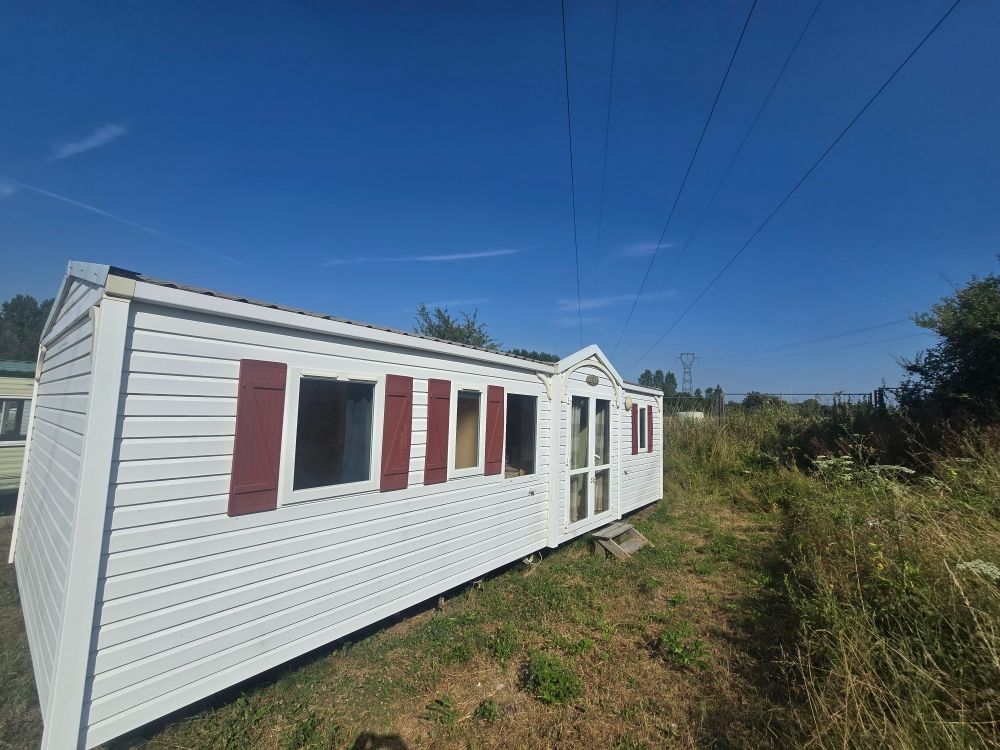 Mobil-home blanc aux volets rouges dans un champ herbeux sous un ciel bleu azur