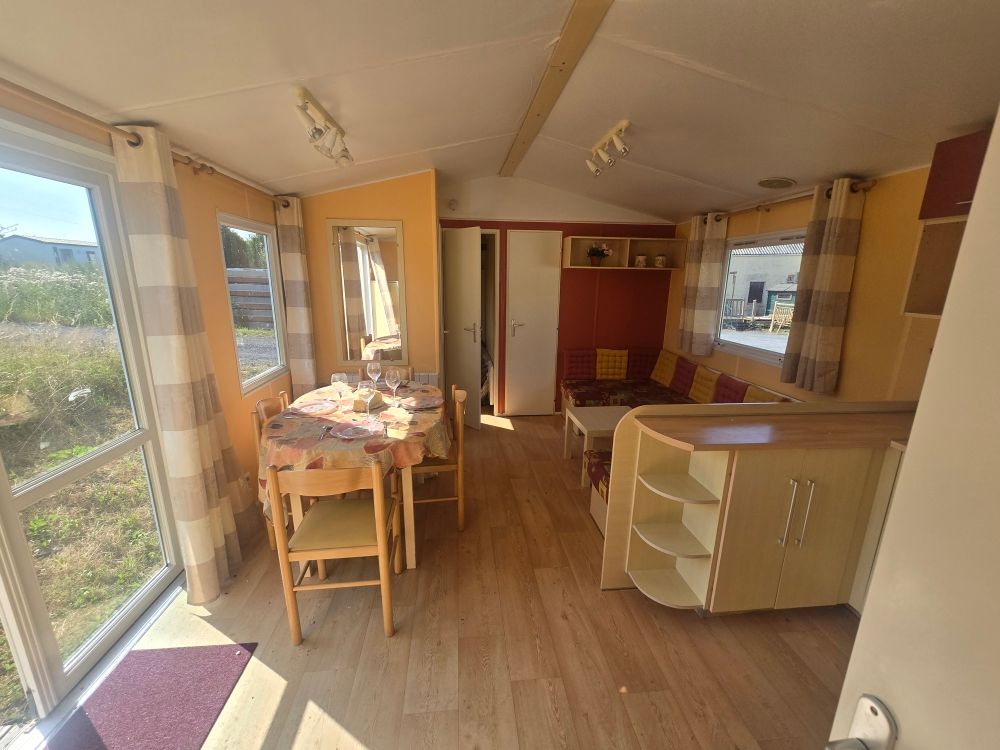 Cuisine et salle à manger lumineuse avec table, chaises et placards en bois, éclairée par de grandes fenêtres et une porte vitrée.