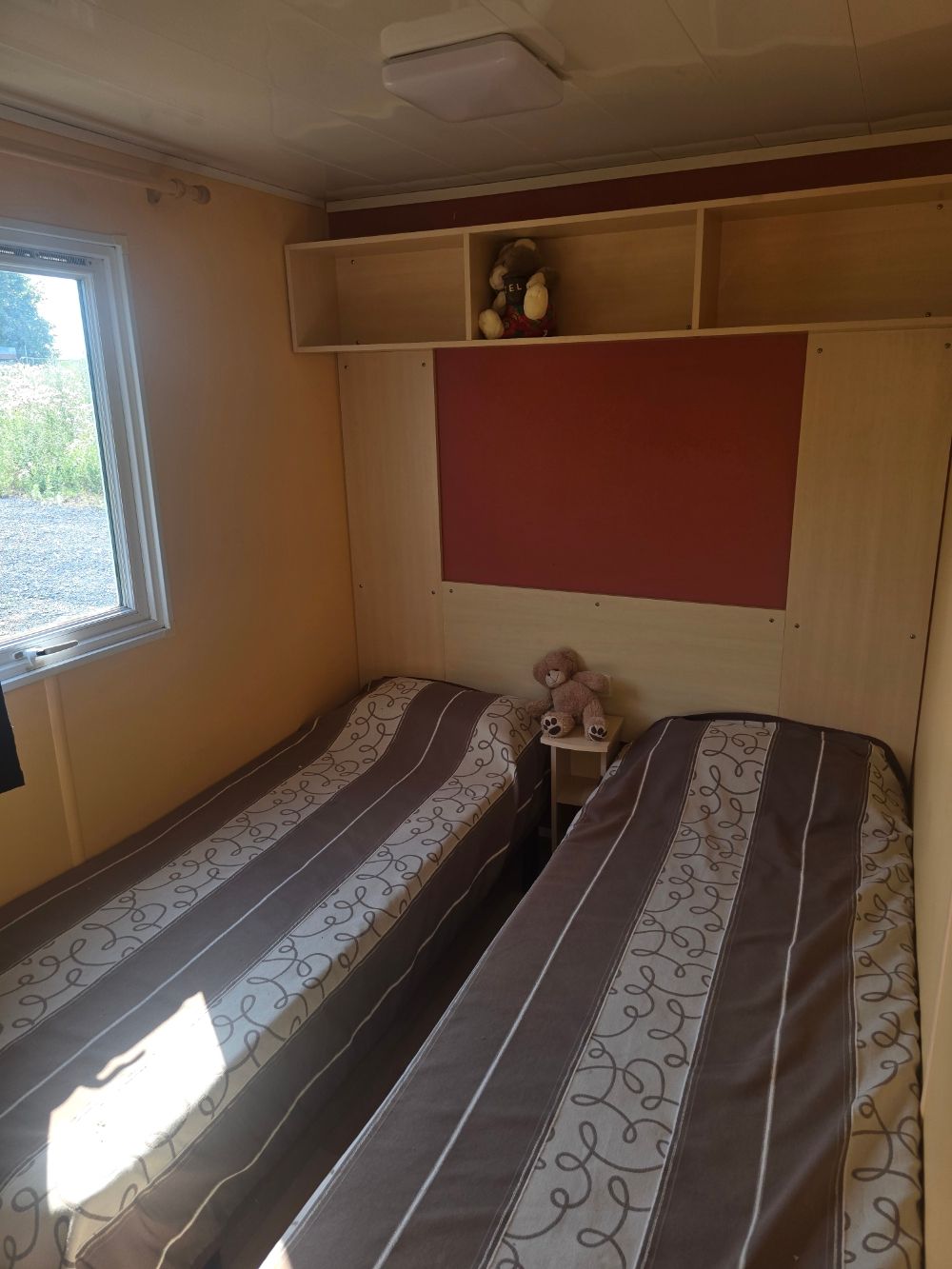 Petite chambre avec deux lits simples, une fenêtre, des étagères murales et un panneau mural rouge
