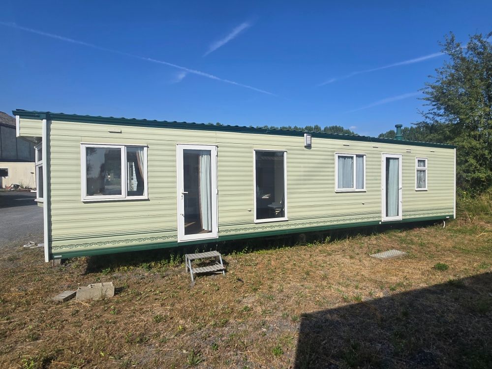 Mobil-home vert clair avec des boiseries et une porte d'entrée blanches, stationné sur un terrain herbeux sous un ciel bleu.
