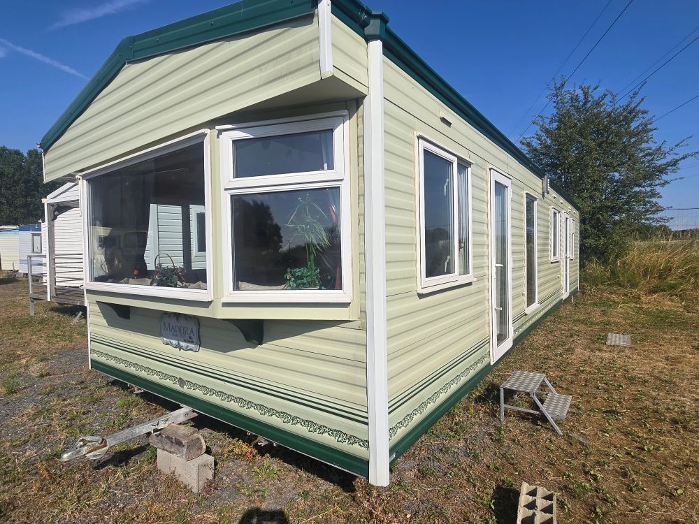 Petite maison mobile beige aux finitions vertes, sur un terrain herbeux sous un ciel bleu.