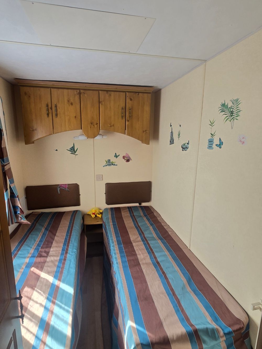 Petite chambre avec deux lits simples, couvre-lits à rayures et armoires murales en bois au-dessus des têtes de lit.