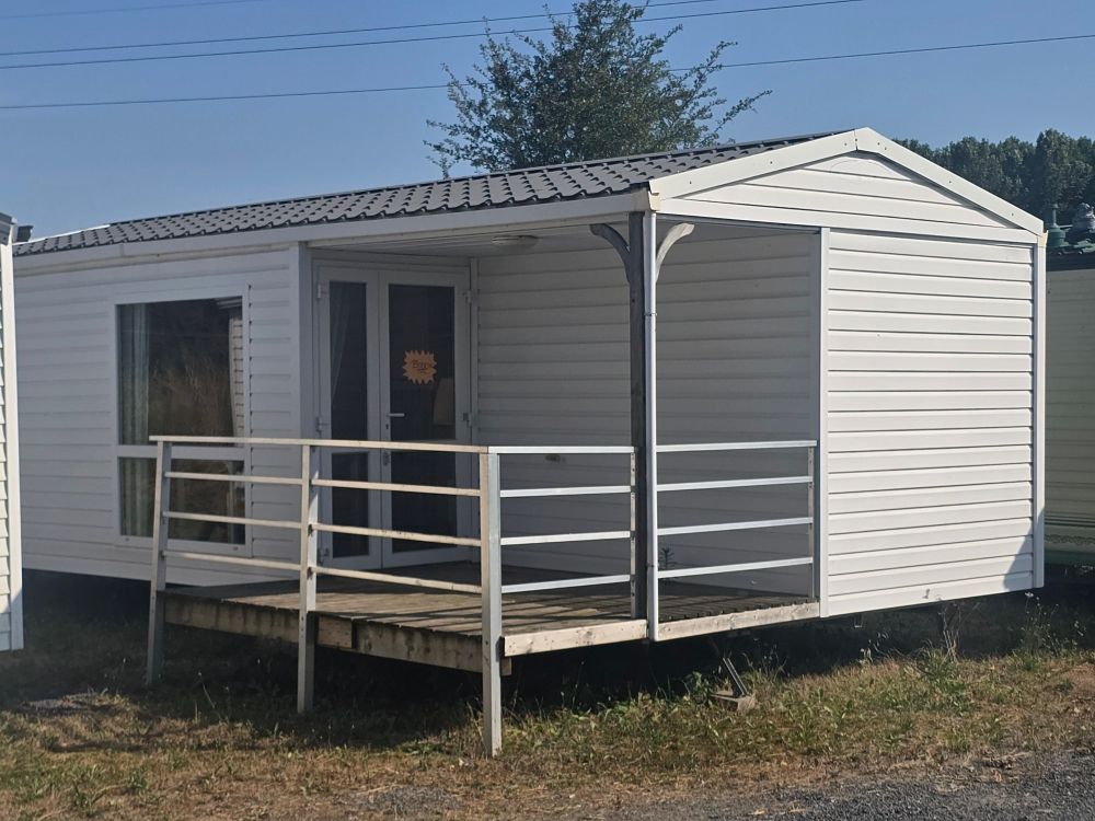 Petite maison mobile blanche avec porche et rampe d'accès, installée sur un terrain herbeux sous un ciel dégagé.