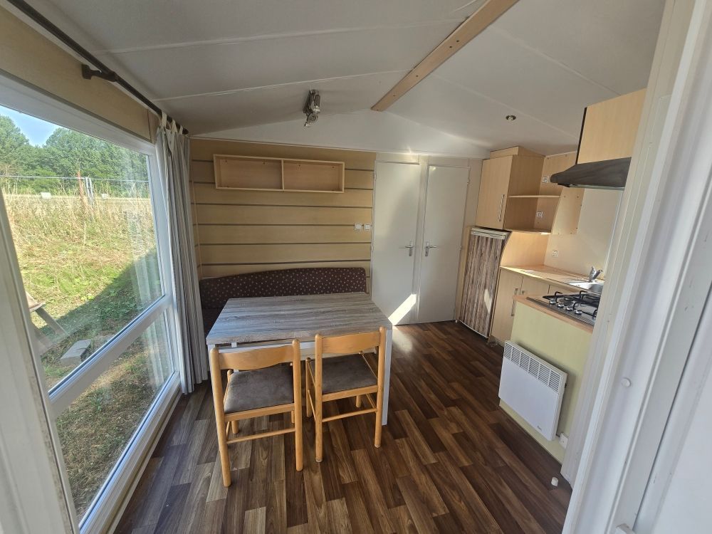 Petite cuisine et coin repas avec parquet, table, chaises, fenêtre ouverte et un lit le long du mur de gauche.