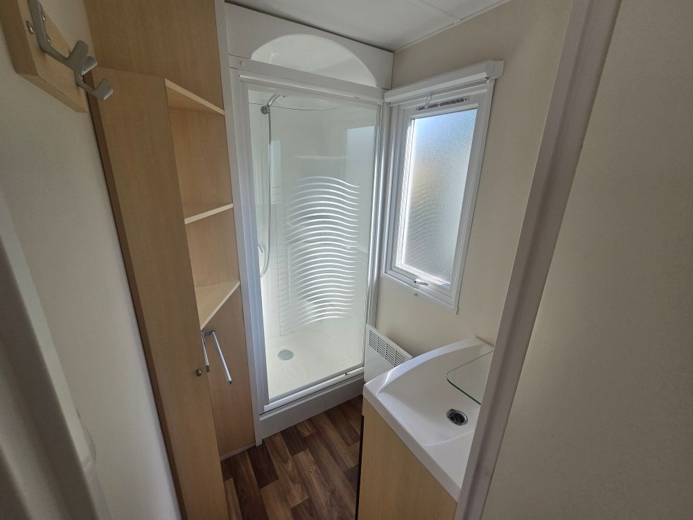 Salle de bain étroite avec cabine de douche, fenêtre dépolie, lavabo et étagères en bois.